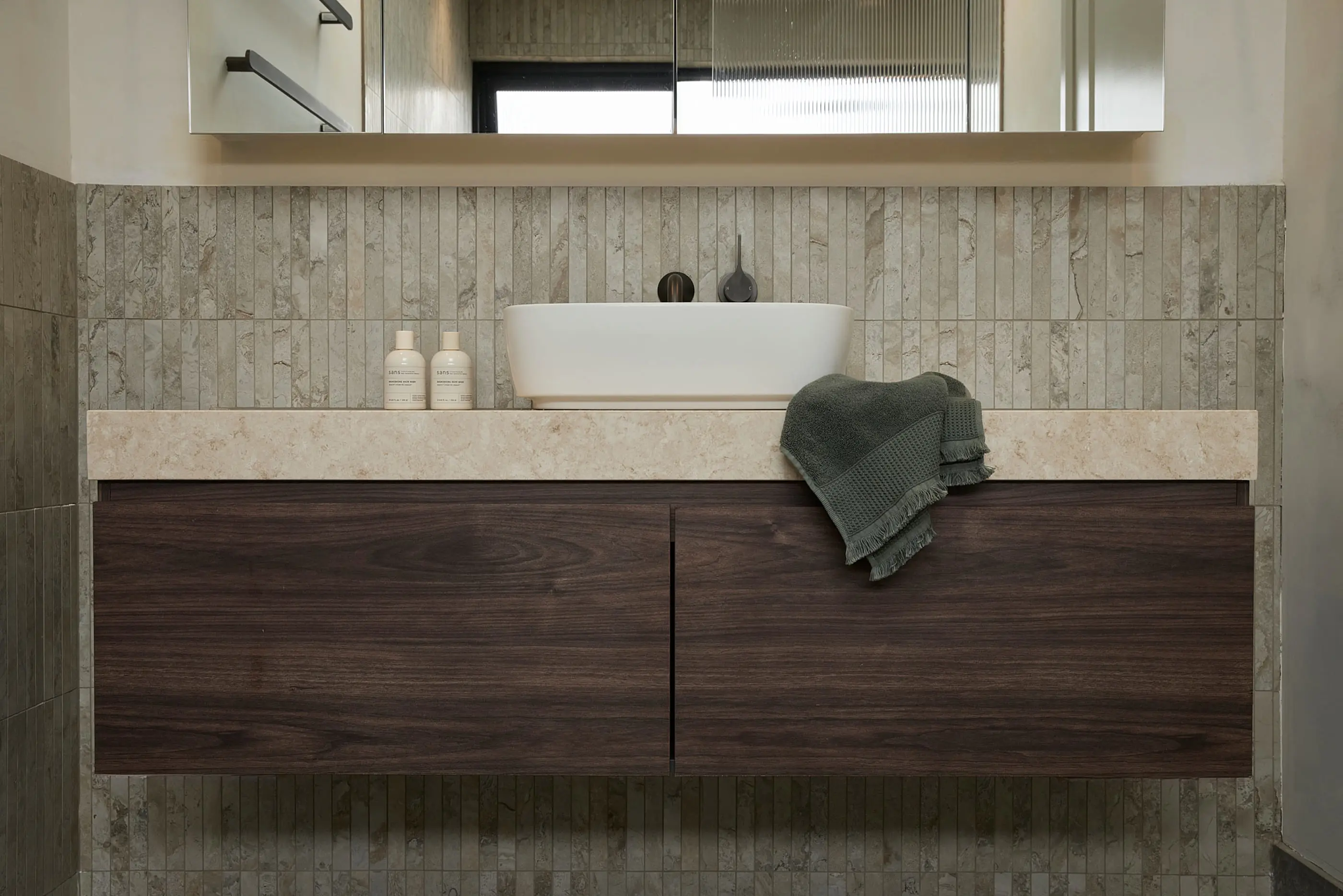 Modern bathroom vanity with white vessel sink, dark wood cabinets, stone countertop, and green towel on marble-tiled wall.