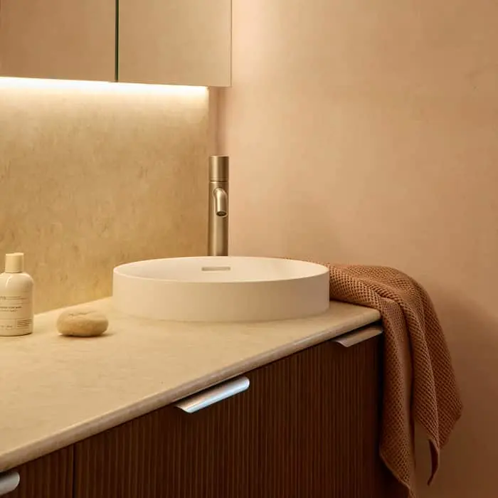 Modern bathroom vanity with round white sink, metallic faucet, beige countertop, and brown towel in warm lighting.