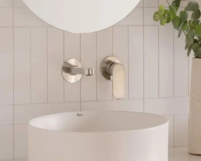 Modern bathroom with white circular sink, wall-mounted brushed nickel faucet, round mirror, and small plant in corner.