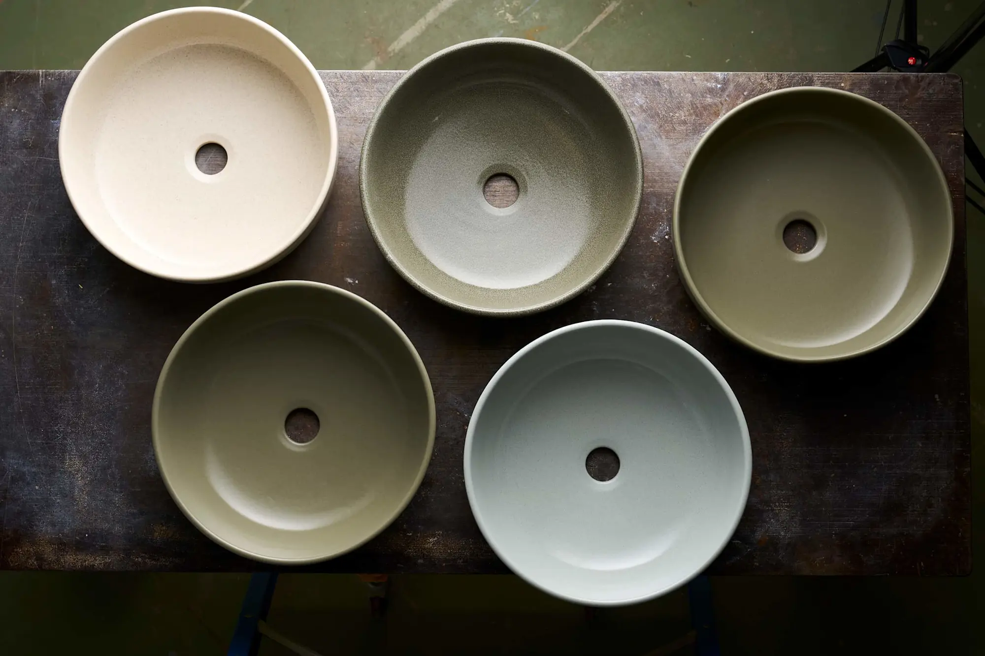 Five ceramic vessel sinks in neutral colors arranged on a dark surface, showing various earth-toned finishes.