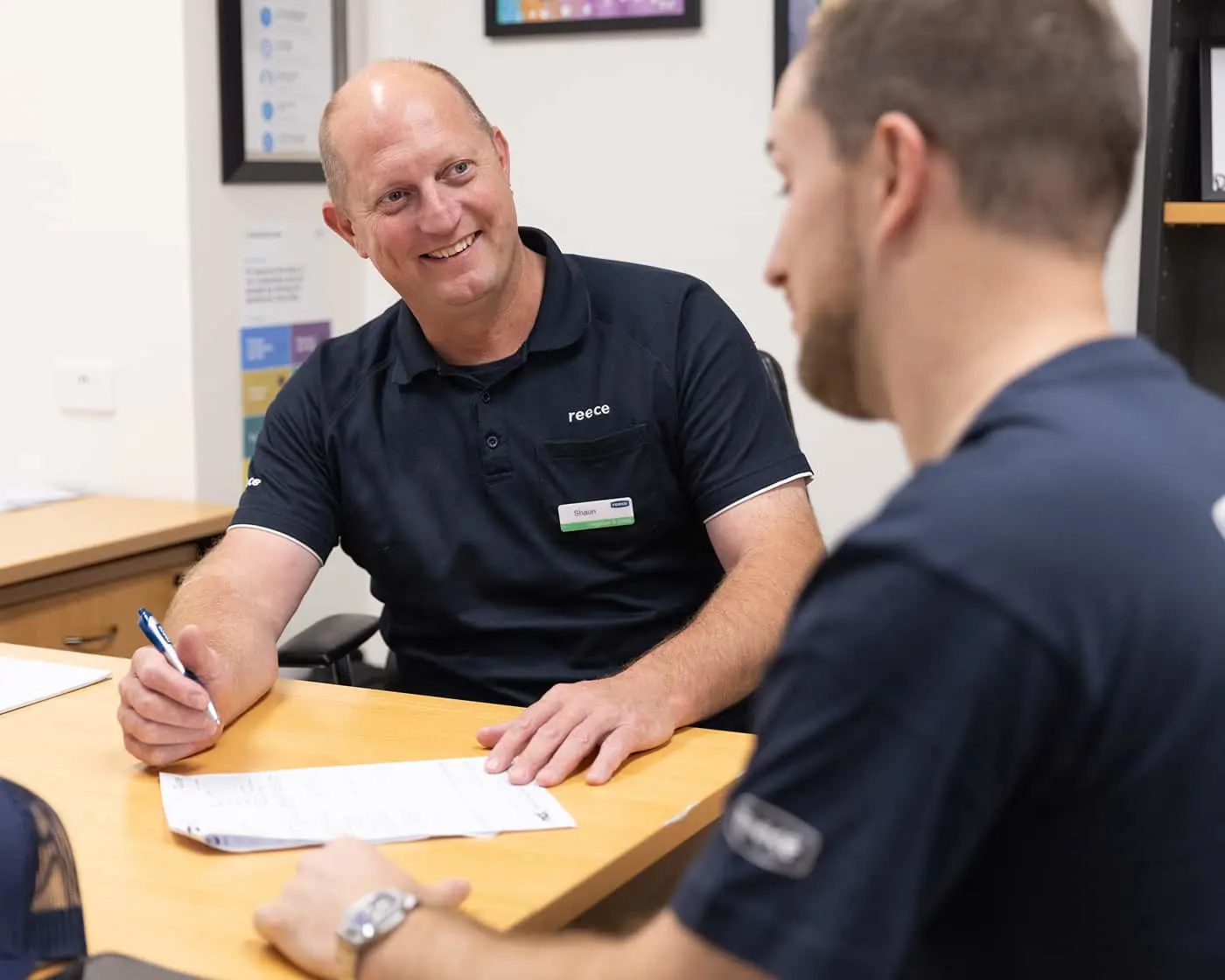Two people in navy polo shirts at a wooden desk, one smiling while writing on paperwork in an office setting.
