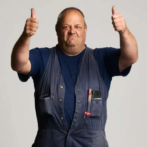 Person in blue work overalls and navy shirt giving thumbs up with both hands against light background.