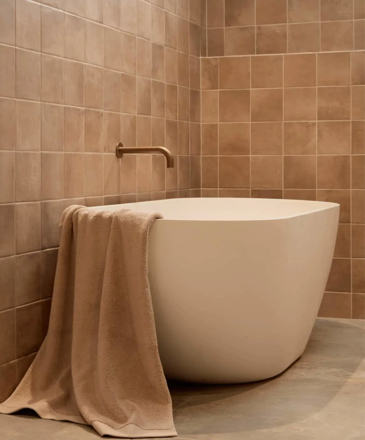 Modern white freestanding bathtub with beige towel against terracotta tiled walls and brass wall-mounted faucet.