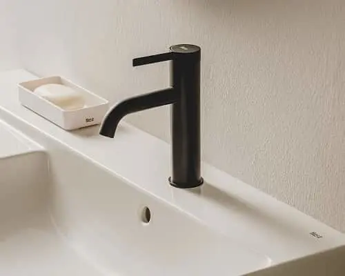 Black cylindrical bathroom faucet mounted on white sink with soap dish in background against beige wall