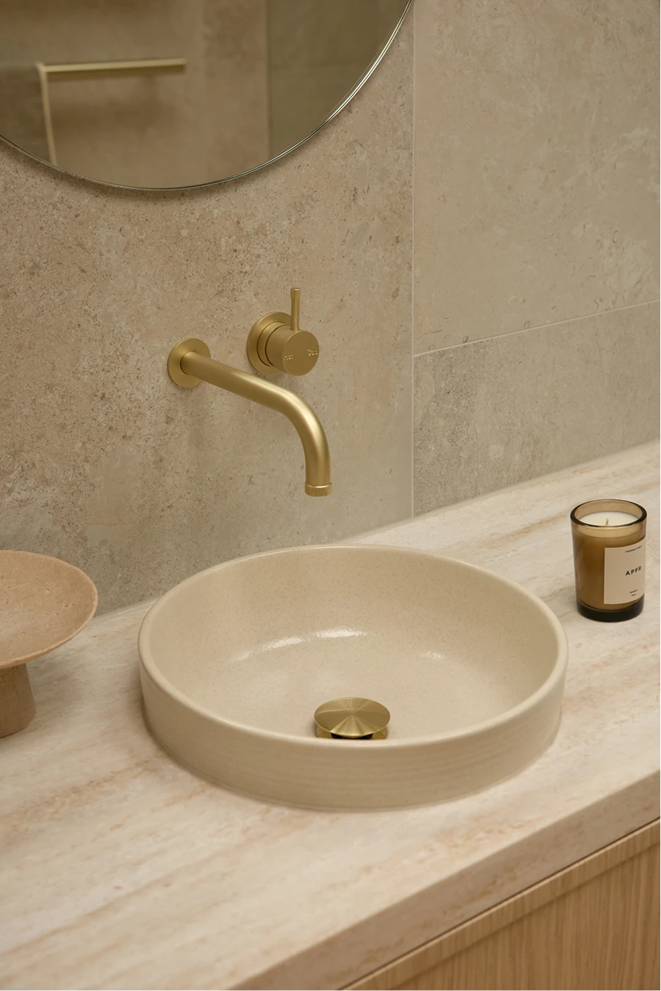 Beige bathroom with round vessel sink, brass wall-mounted faucet, round mirror, and marble countertop.