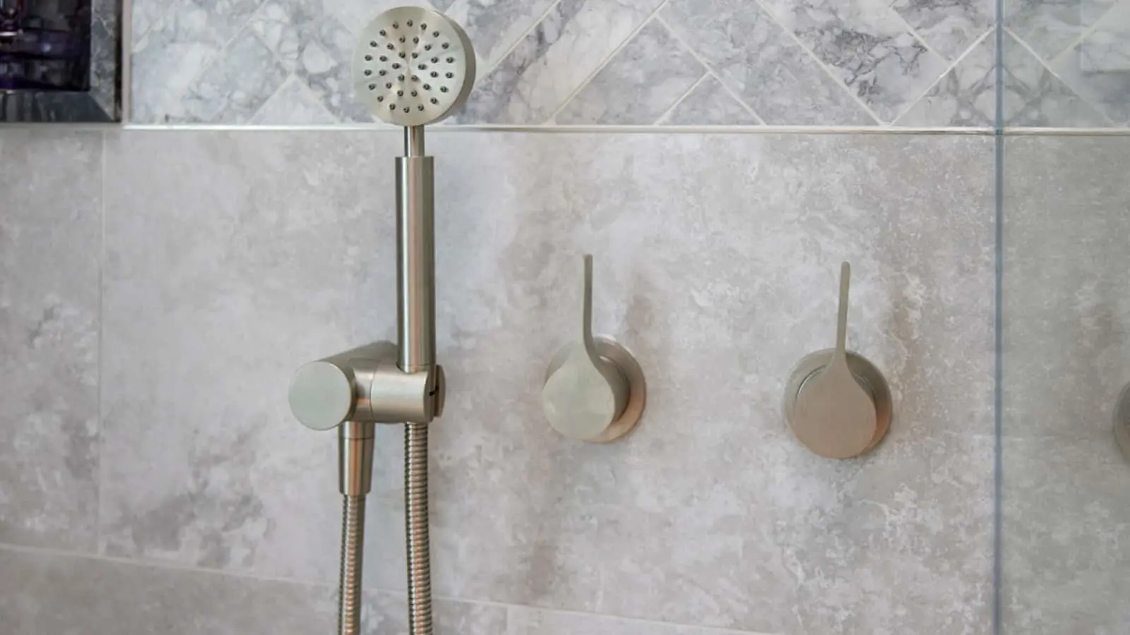 Modern brushed nickel handheld shower head with flexible hose mounted on marble-tiled bathroom wall with two control knobs.