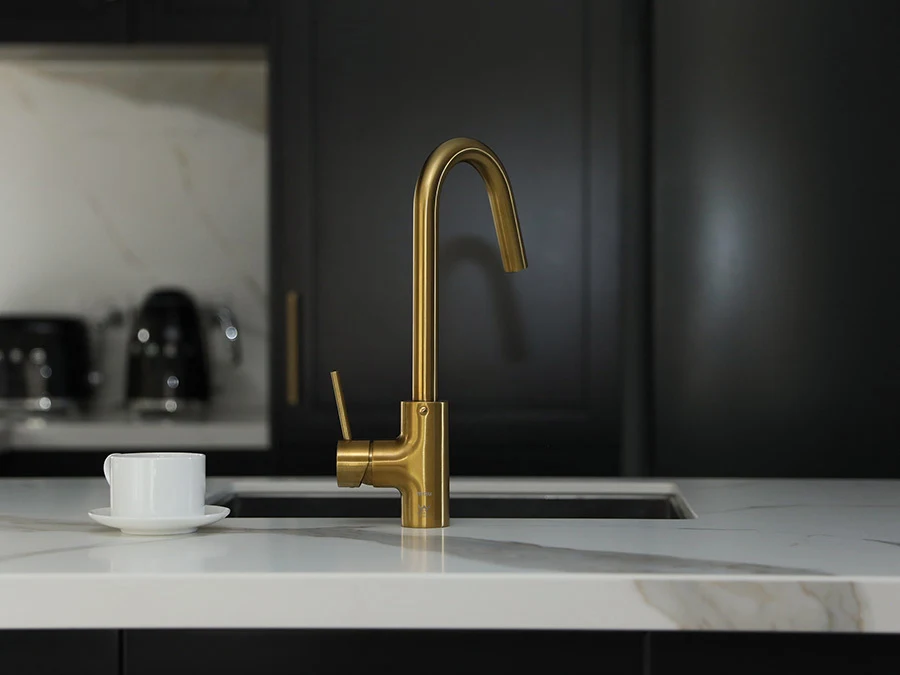 Modern brass kitchen faucet on white marble countertop with sink and white coffee cup, against dark cabinetry.