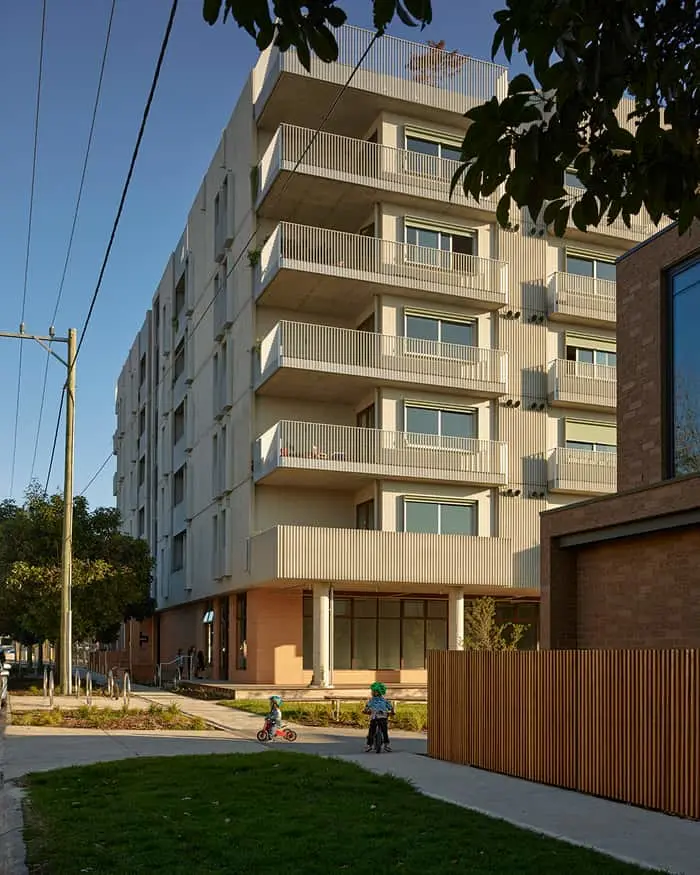 Modern apartment building with white balconies and ground-floor retail space, children playing on bikes in front.