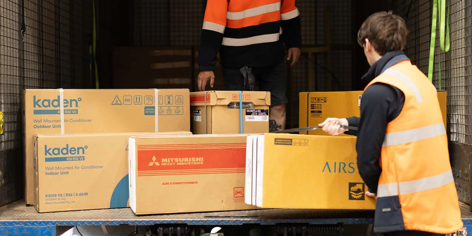 Two workers in reflective vests handling boxed air conditioner units on a truck.