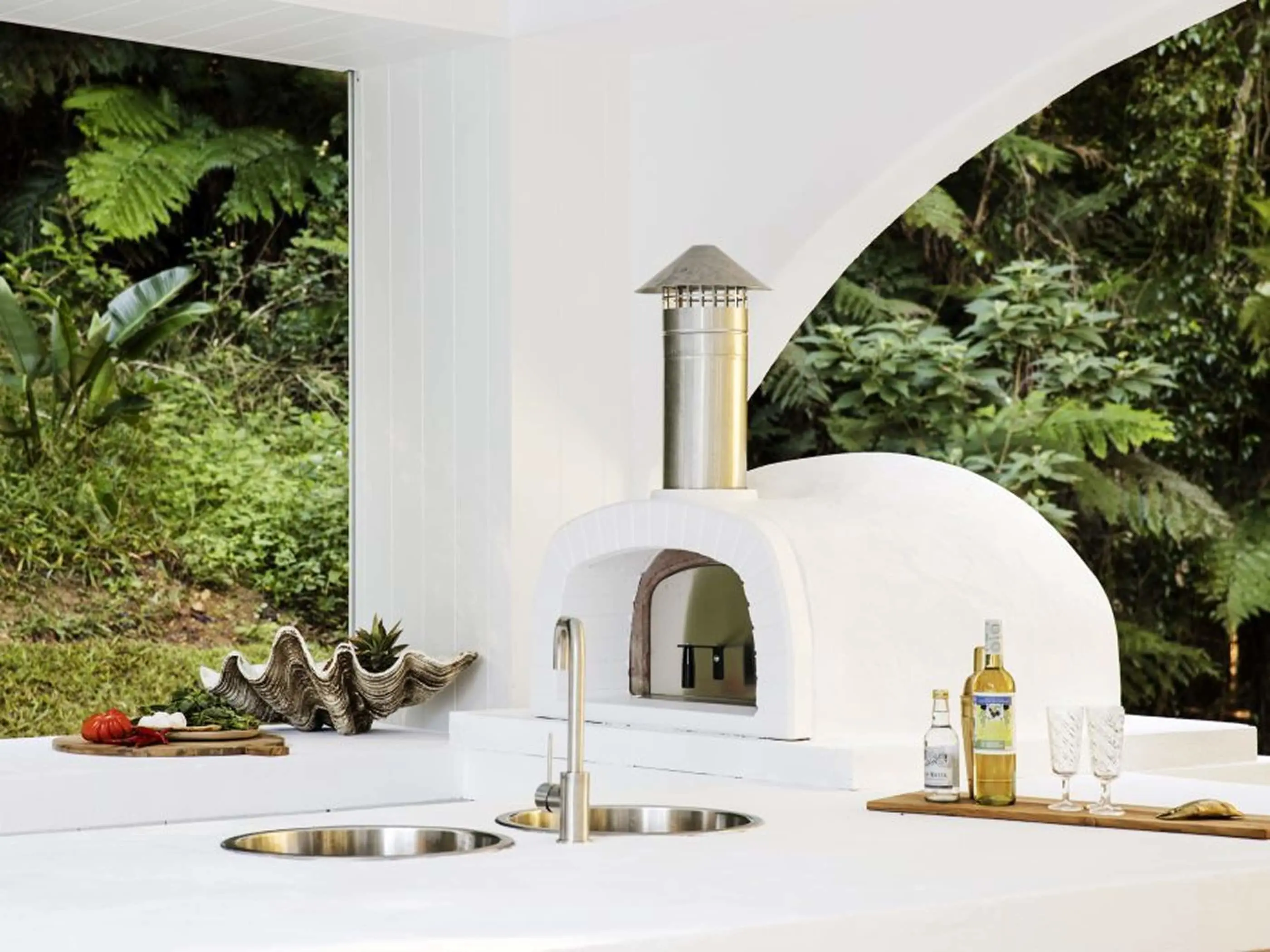 White outdoor pizza oven with stainless steel chimney on counter with sink, drinks, and tropical foliage backdrop.