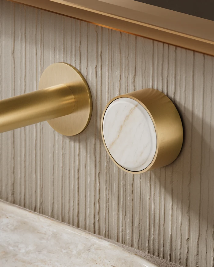 Brushed gold wall-mounted faucet spout and round control knob with marble insert on a textured cream stone wall.