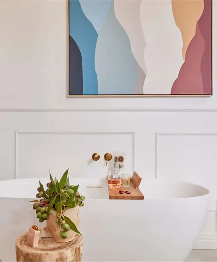Modern white bathroom with freestanding tub, wooden bath caddy, plant on stump, and colorful abstract artwork on wall.