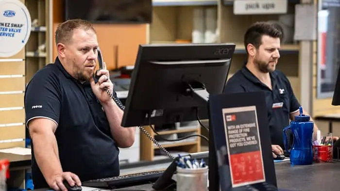 Two staff members working at a service counter with computers, one speaking on a telephone.