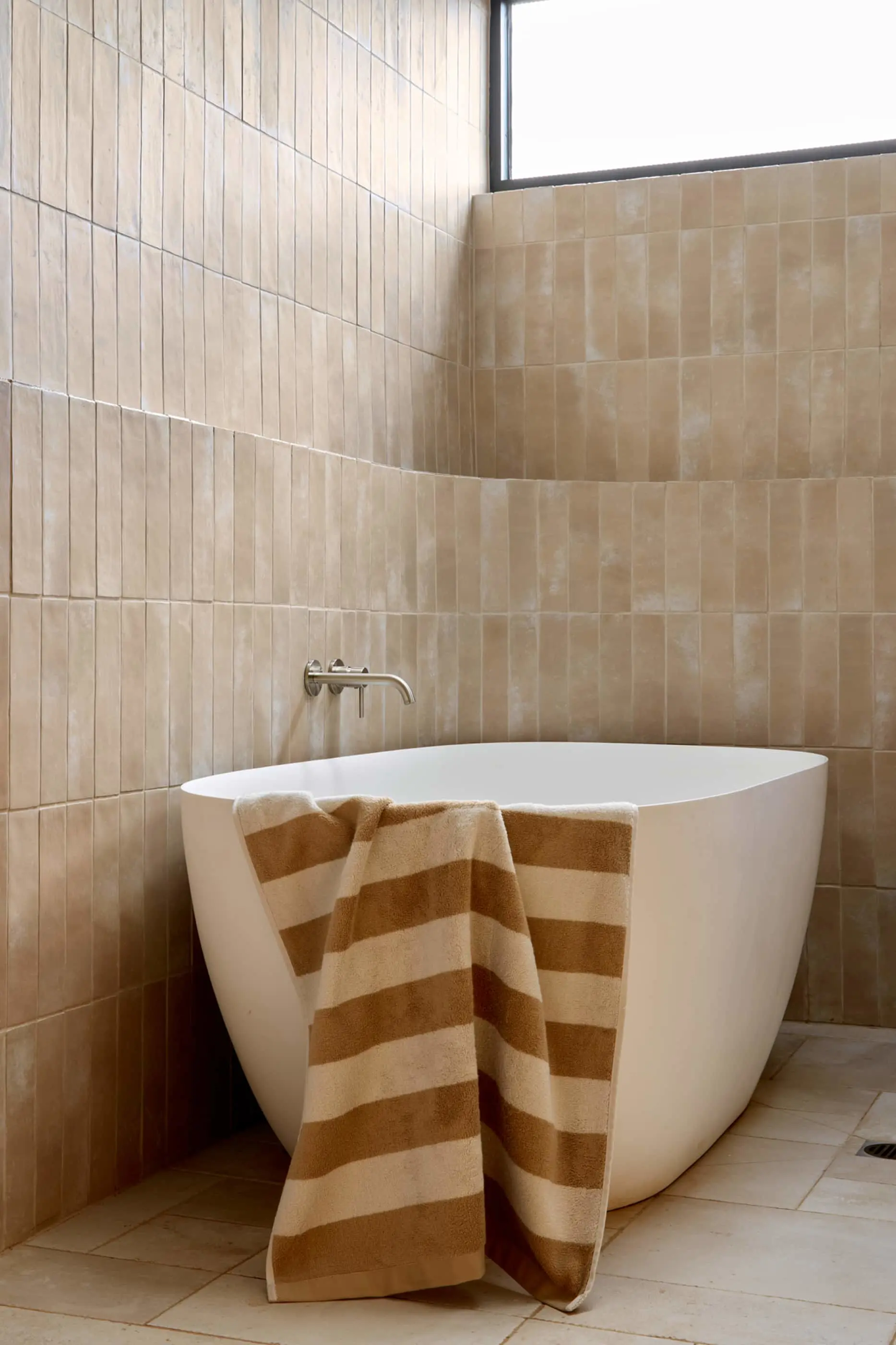 Modern white freestanding bathtub with brown striped towel in a bathroom with beige vertical tile walls and window.