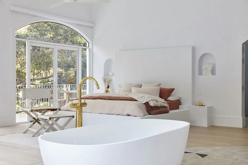 Bright bedroom with white freestanding bathtub, gold faucet, white bed with rust-colored linens, and arched windows overlooking trees.