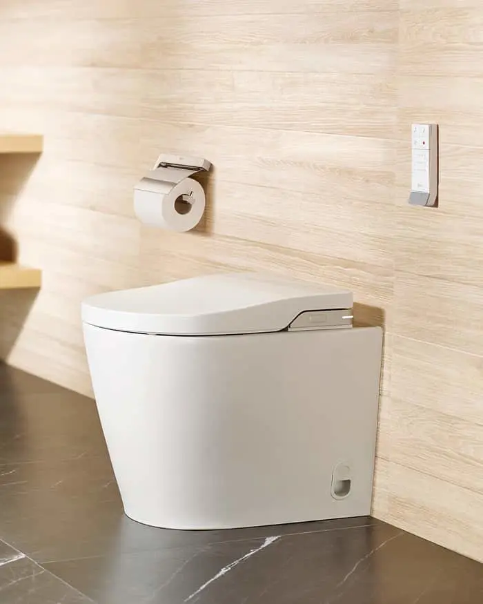 Modern white toilet with closed lid against light wood wall panels, toilet paper holder and control panel mounted nearby.