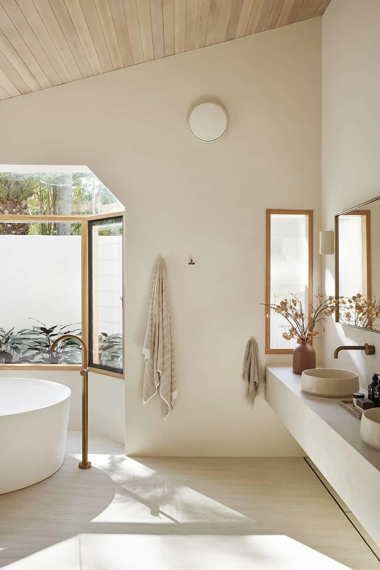 Modern bathroom with white freestanding tub, cream walls, wooden ceiling, large windows, and minimalist sink area with dried flowers.