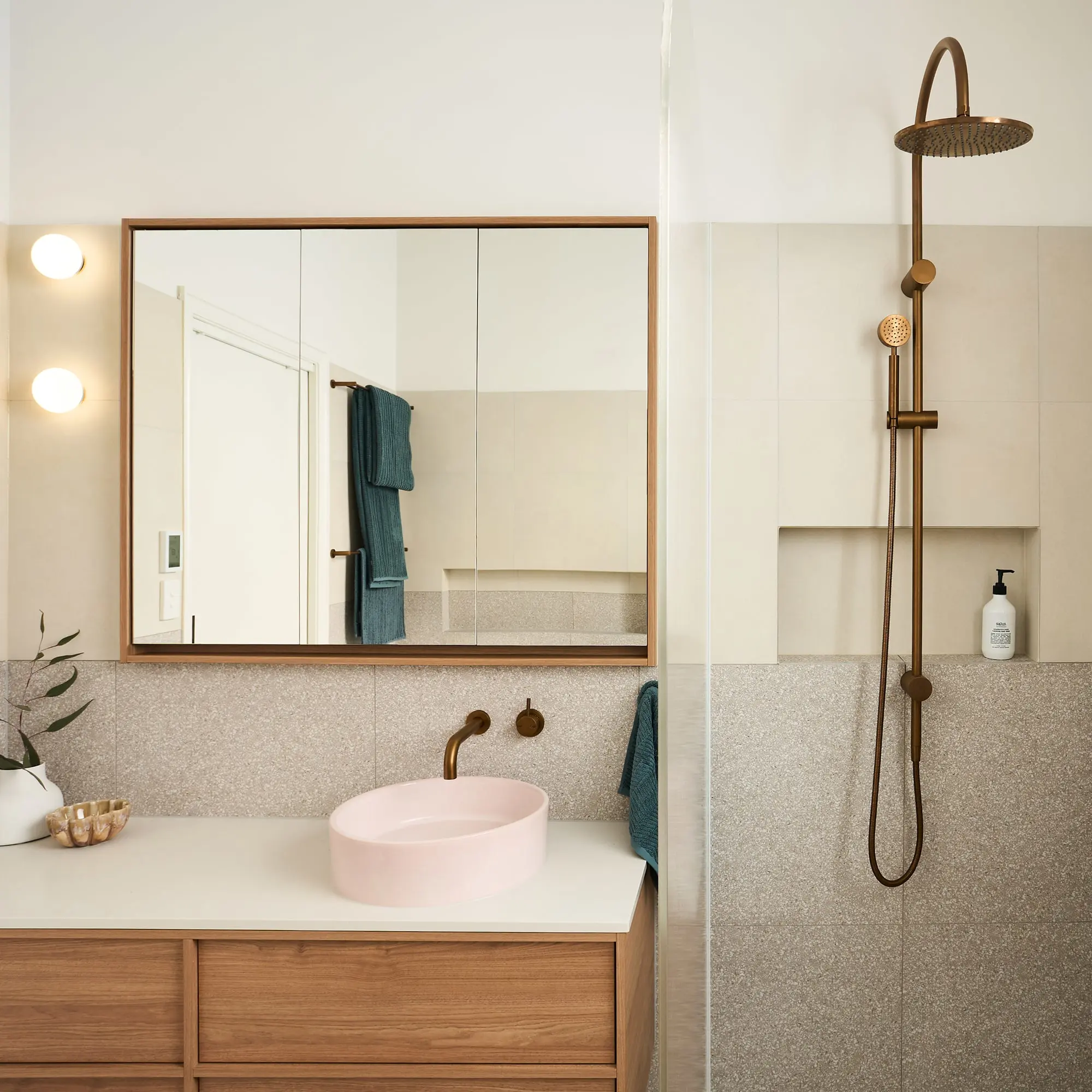 Modern bathroom with pink basin, wooden vanity, large mirror, brass fixtures, and shower with bronze rainfall showerhead.