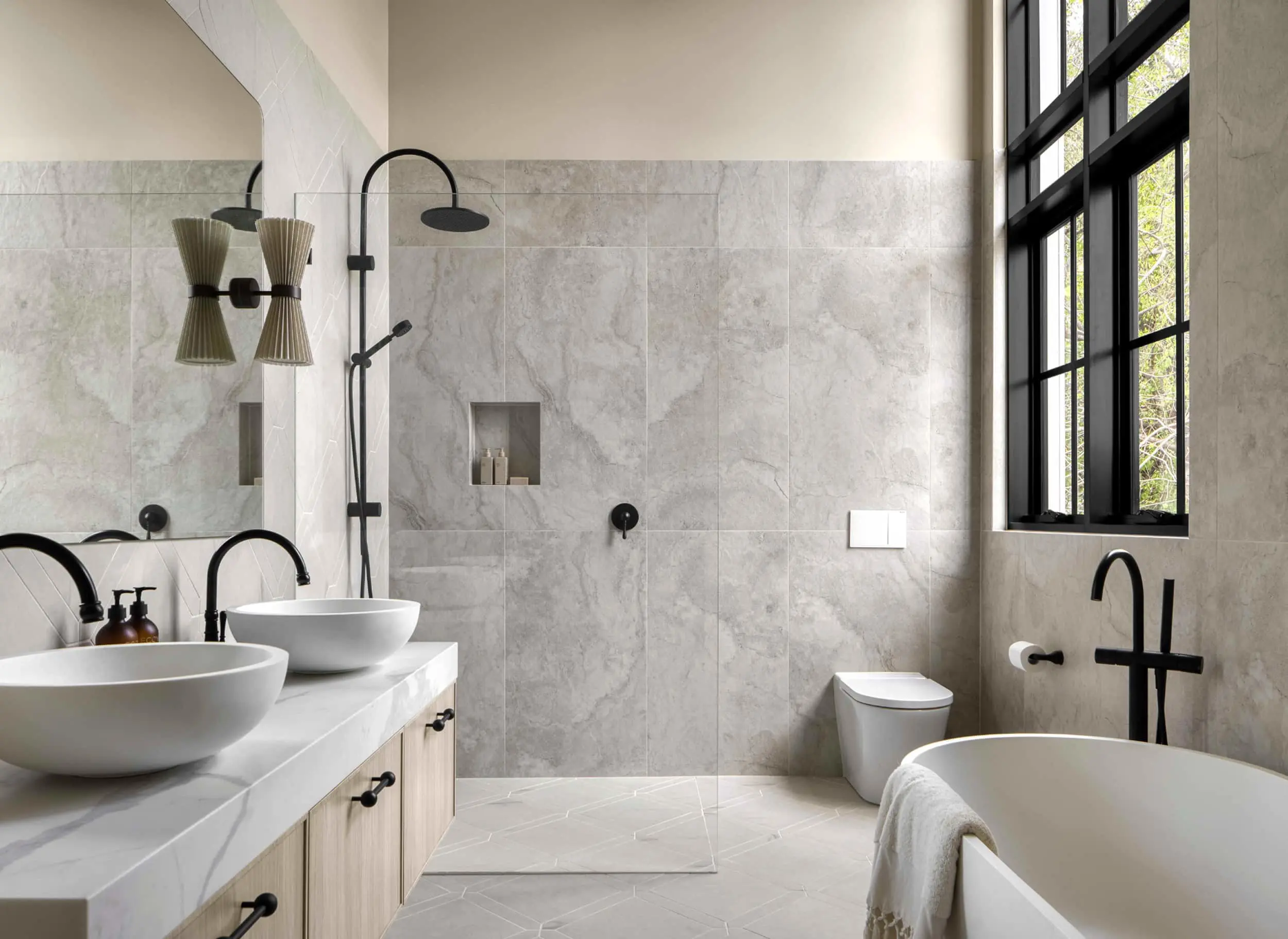 Modern bathroom with marble walls, dual vessel sinks, freestanding tub, black fixtures, and large windows letting in natural light.