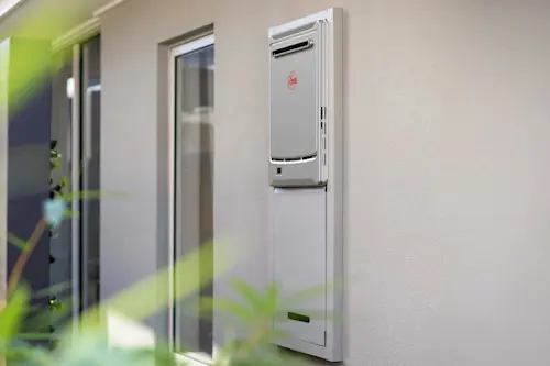 Silver Rheem tankless water heater mounted on white exterior wall next to windows with green plants in foreground.