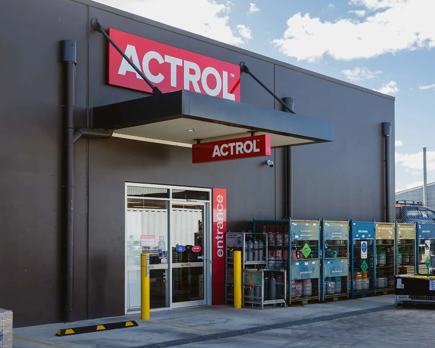 Actrol store exterior with gray building, red signage, glass entrance doors, and storage cages containing industrial supplies.