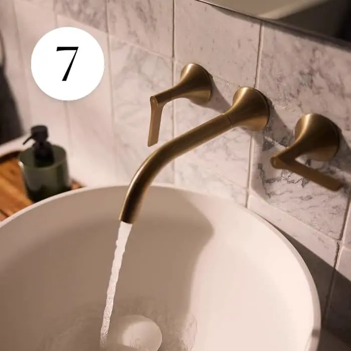 Gold wall-mounted bathroom faucet running water into a white basin, marble tiles background, number 7 overlay.
