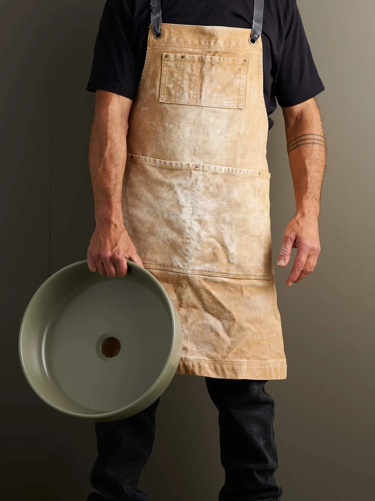 Person wearing tan canvas apron and black shirt holding a light green ceramic vessel with center hole.