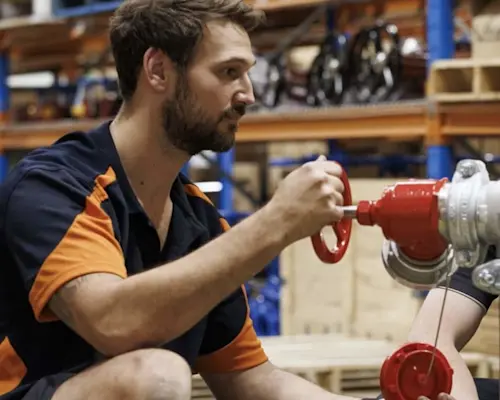 Person in navy and orange uniform working on a red valve component in a warehouse setting.