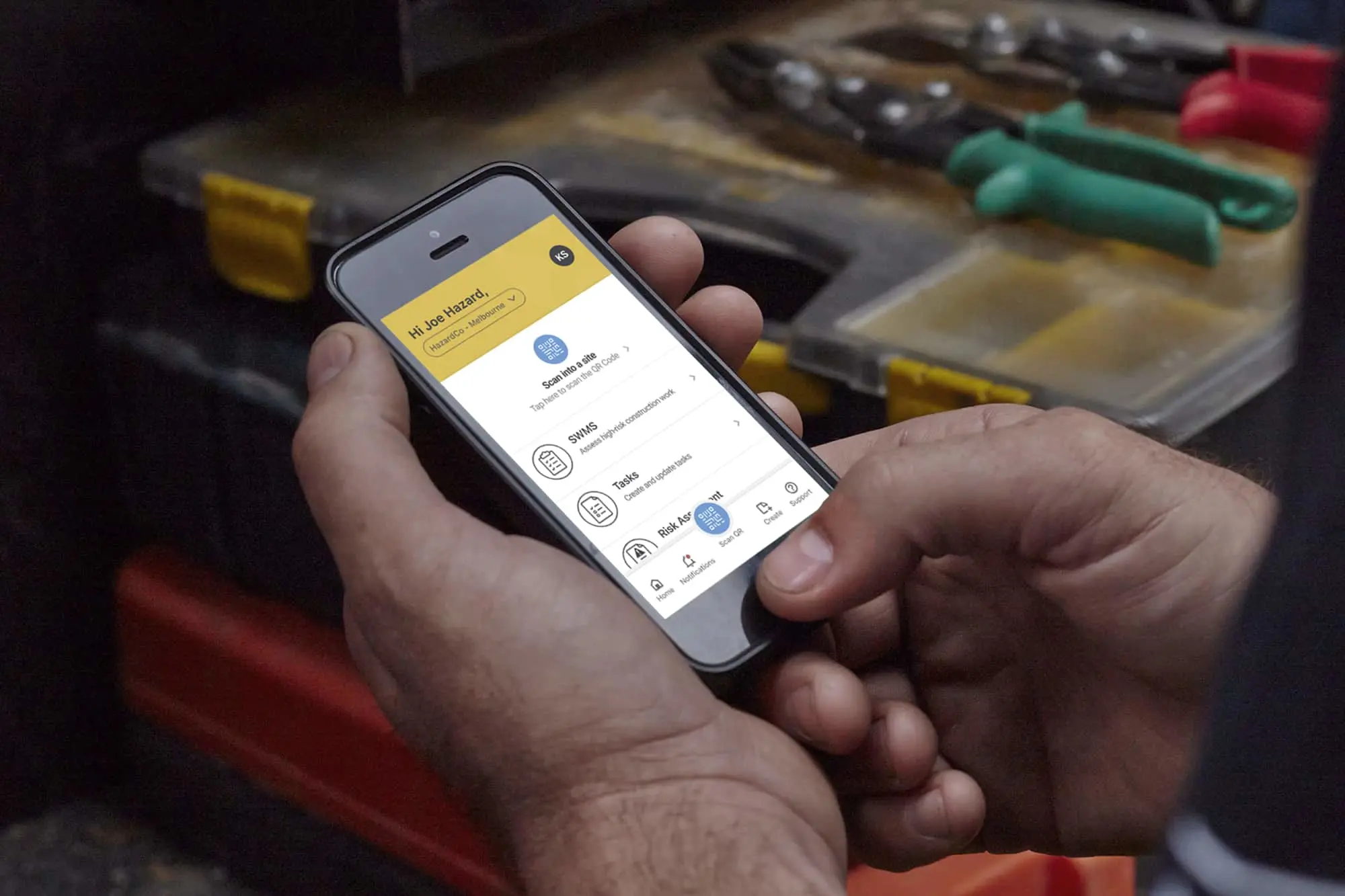 Hands holding smartphone displaying safety app with yellow header, with tools visible in background workspace.