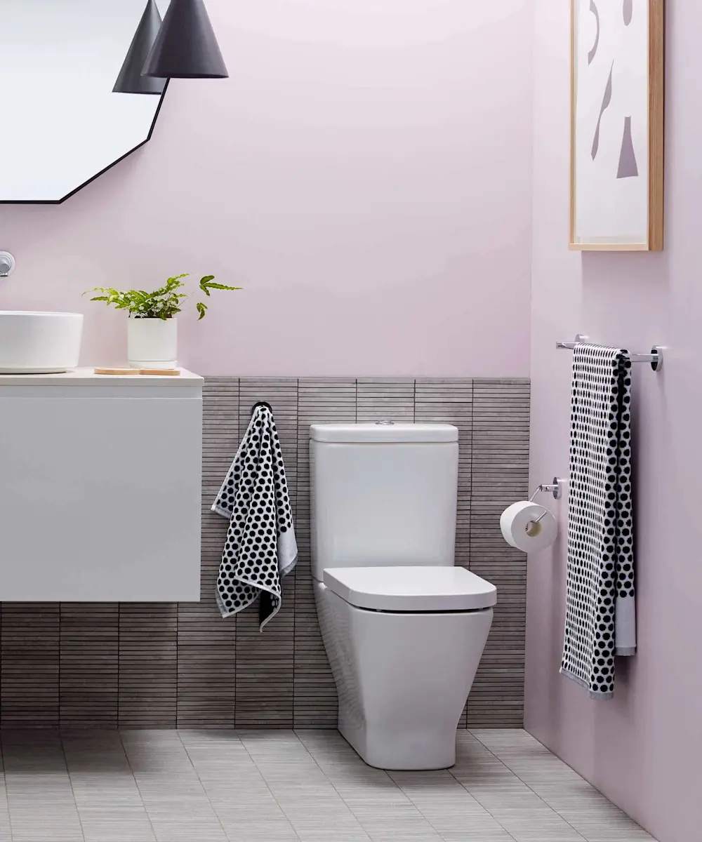 Modern bathroom with pink walls, white toilet, gray tile backsplash, black pendant light, and polka dot towels.