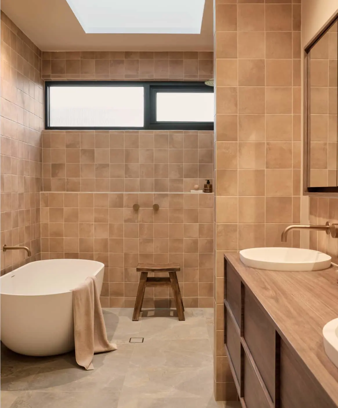 Modern bathroom with beige tiles, wooden vanity with white vessel sink, freestanding bathtub, and wall-mounted faucets.
