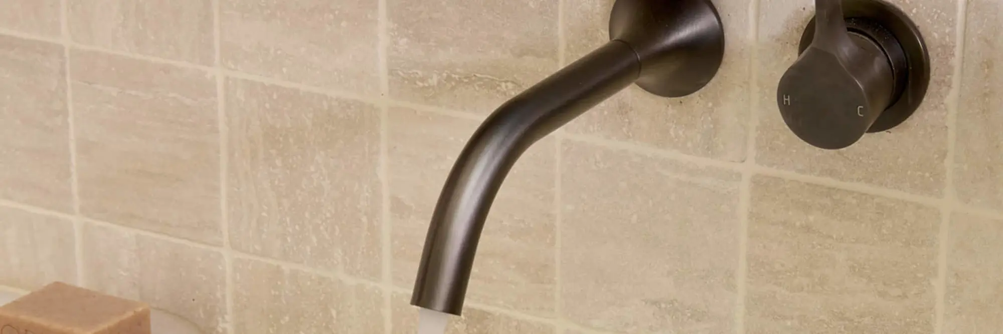 Modern gunmetal wall-mounted bathroom faucet with curved spout against beige tiled wall, control knob visible.