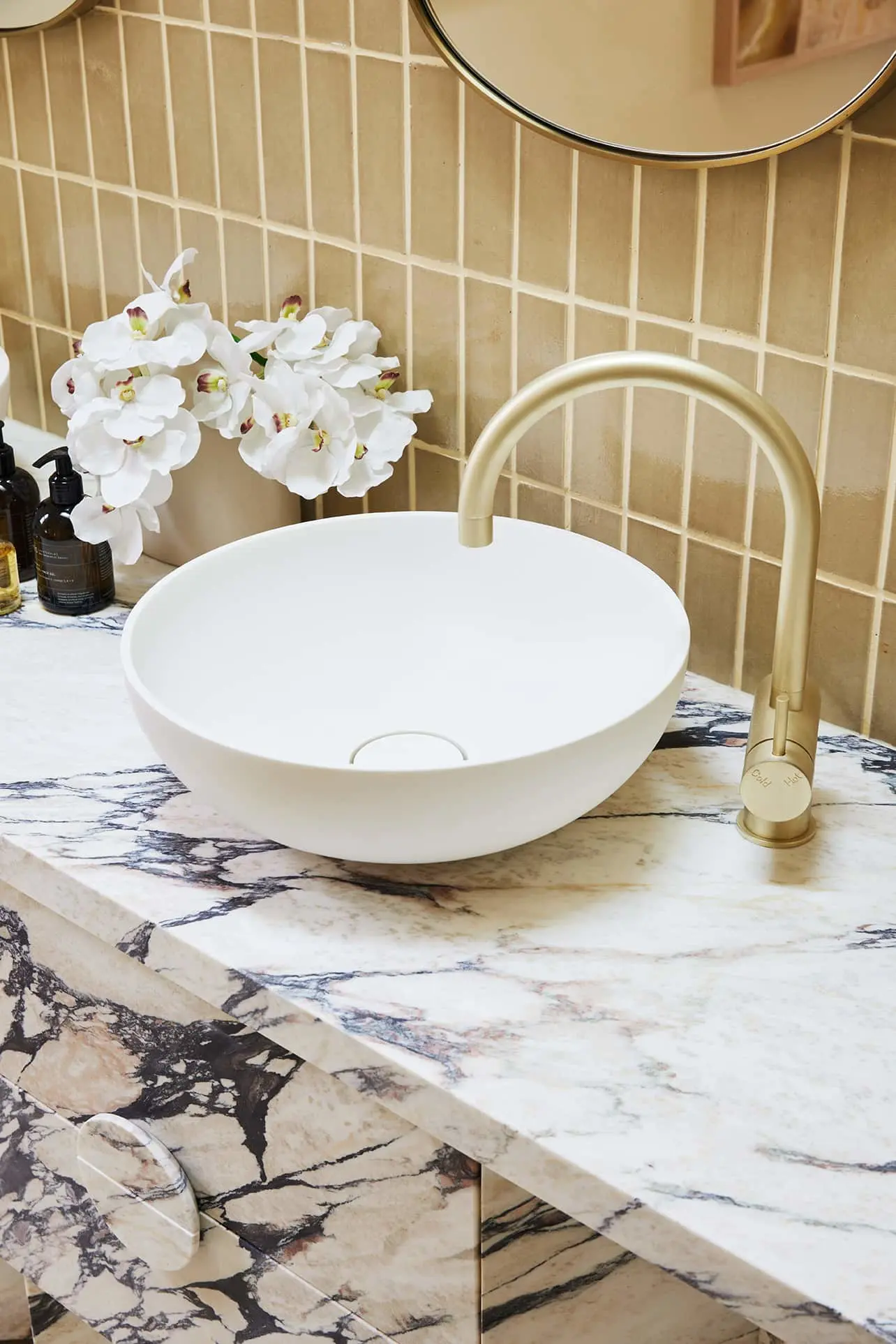 Modern bathroom with white vessel sink, gold faucet, marble countertop, beige tile backsplash, and white orchids.