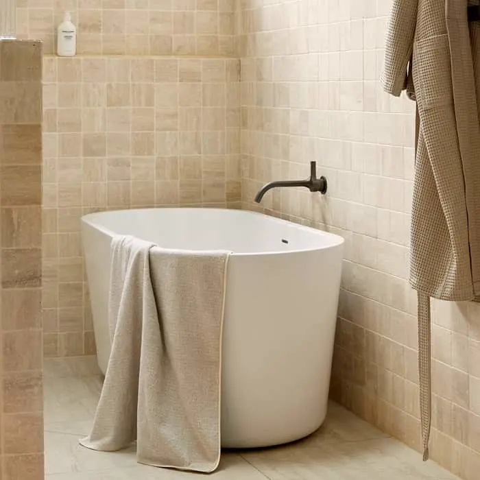 Modern white soaking tub with beige towel in a neutral-tiled bathroom with wall-mounted faucet and hanging robe.
