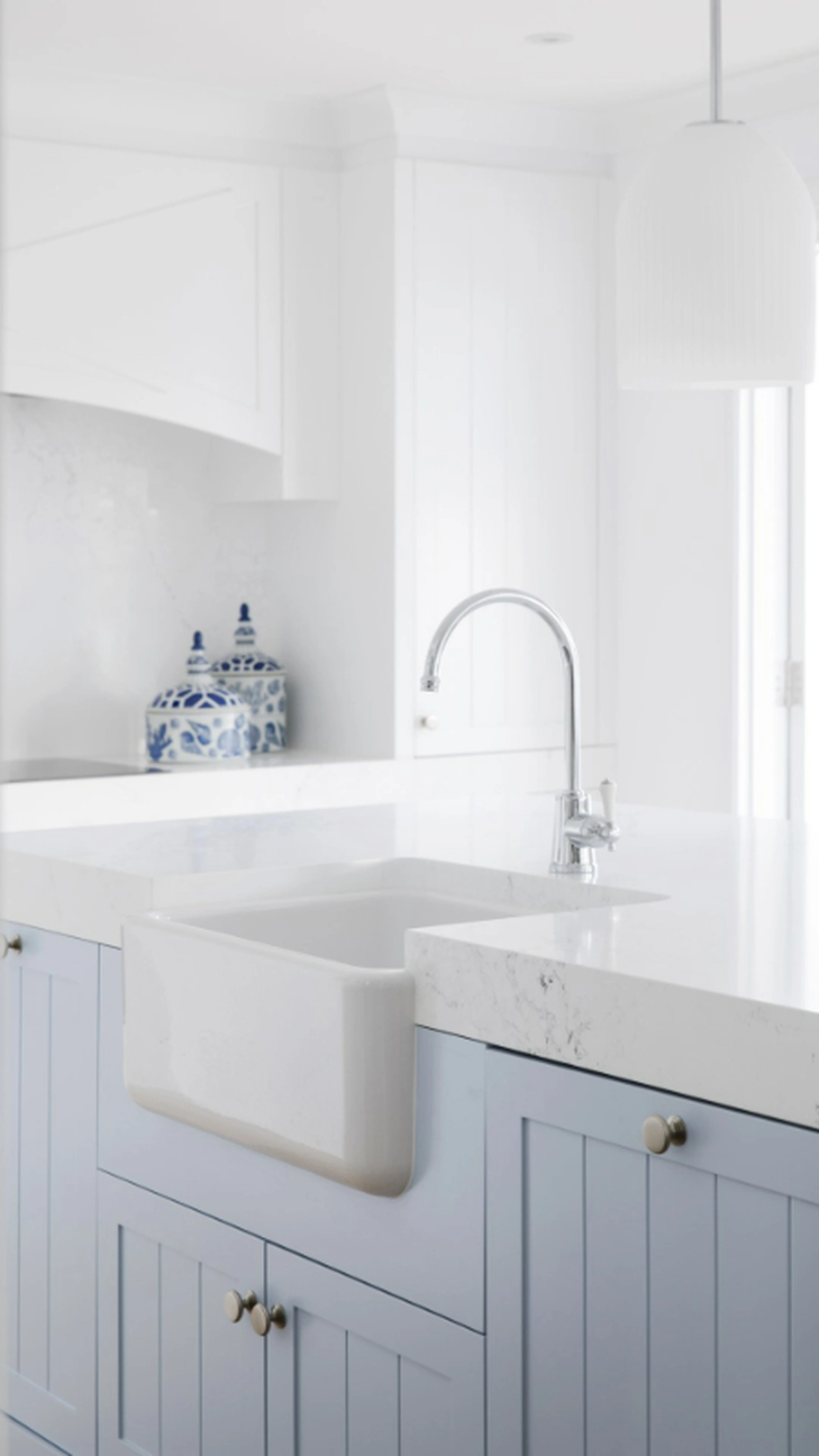 Modern white kitchen with farmhouse sink, chrome faucet, light blue cabinets, and blue-white decorative jar on counter.