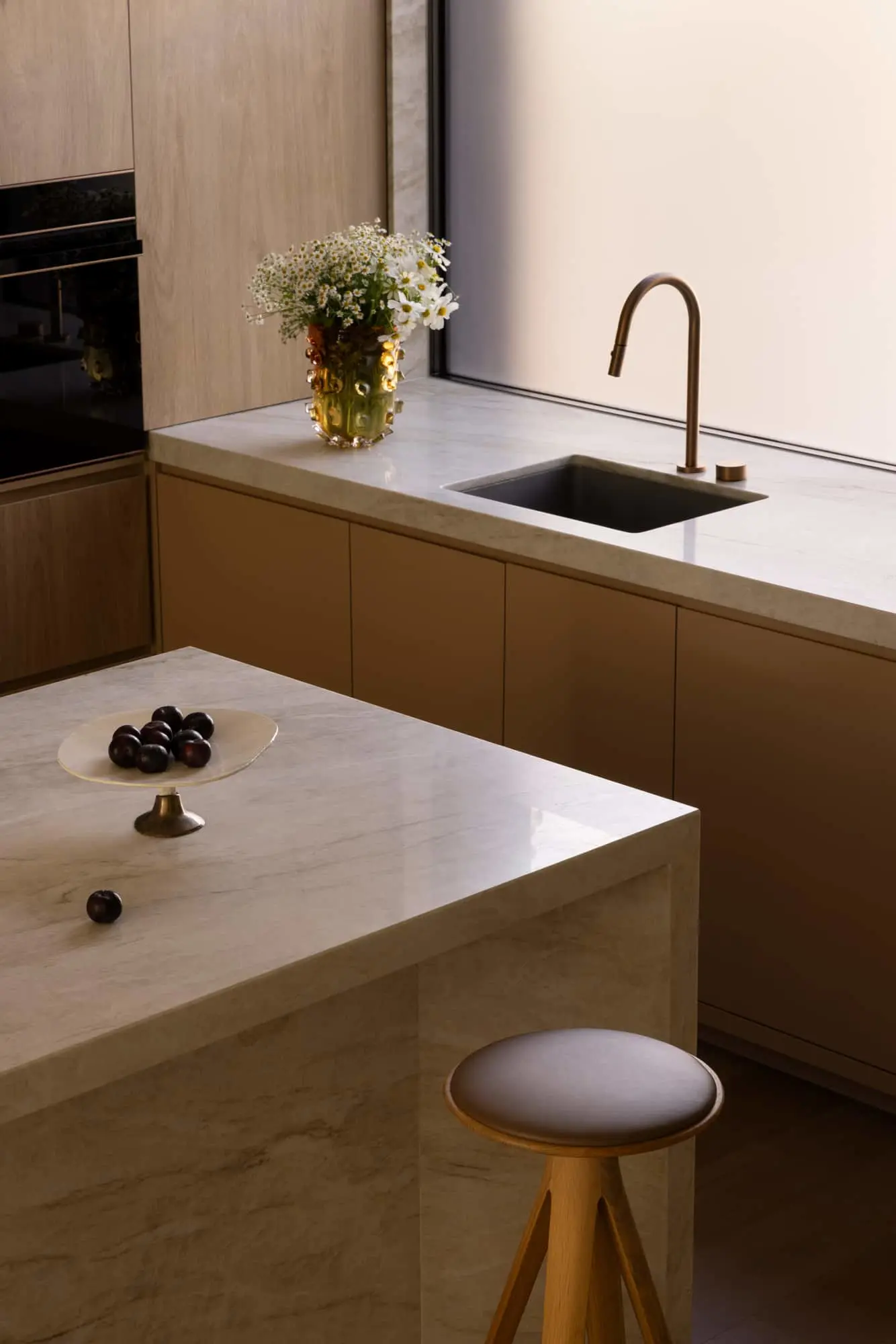 Modern kitchen with marble countertops, brass faucet, white daisies in amber vase, and plums on a pedestal dish.