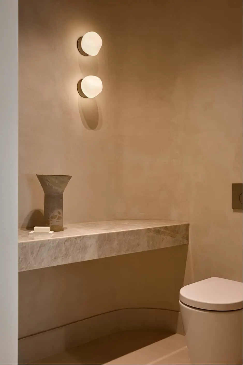 A bathroom with beige plaster walls, a floating marble shelf with a ceramic vase, a double globe wall sconce, and a white toilet.