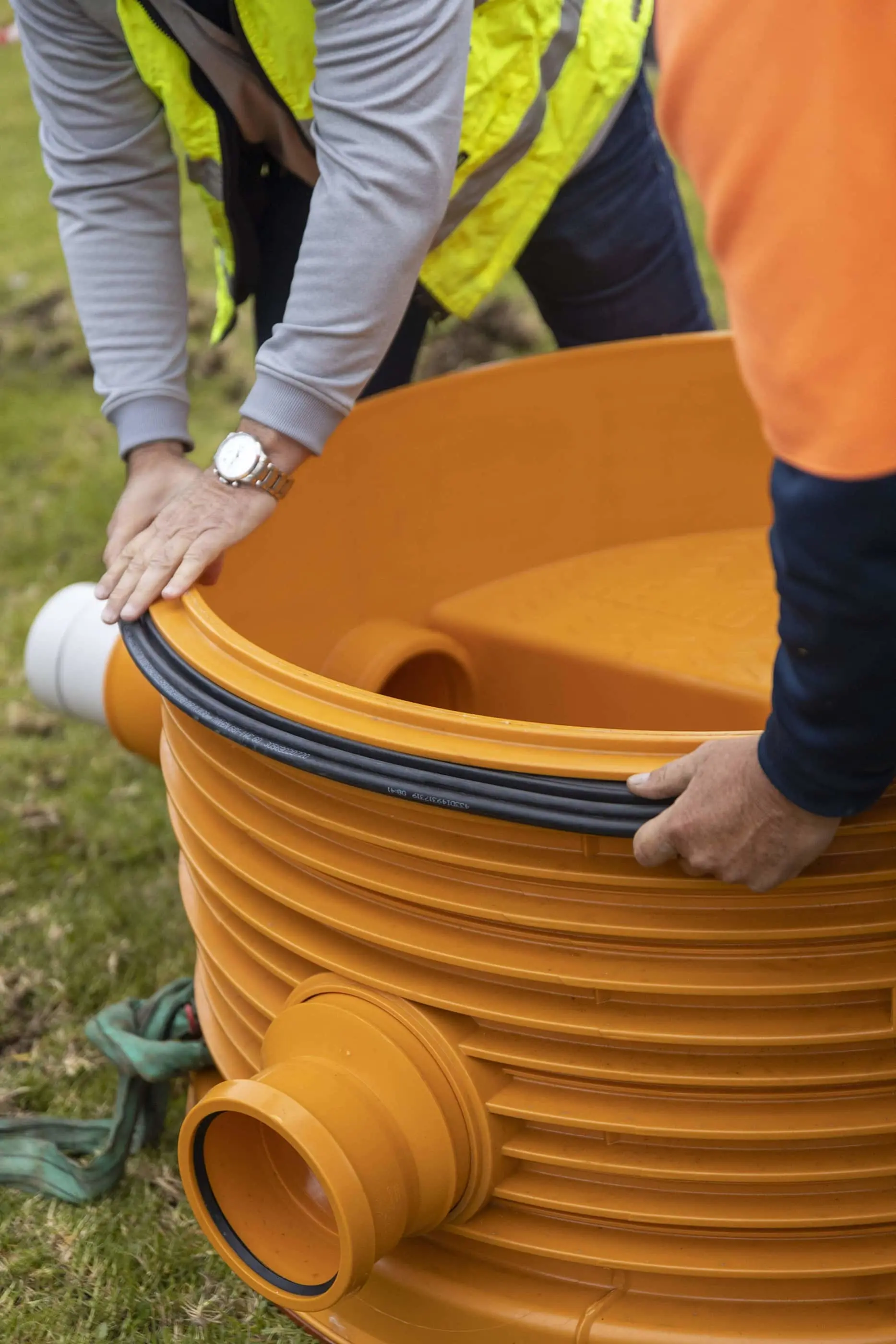 Workers in high-visibility vests installing an orange drainage chamber or inspection pit on grassy ground.