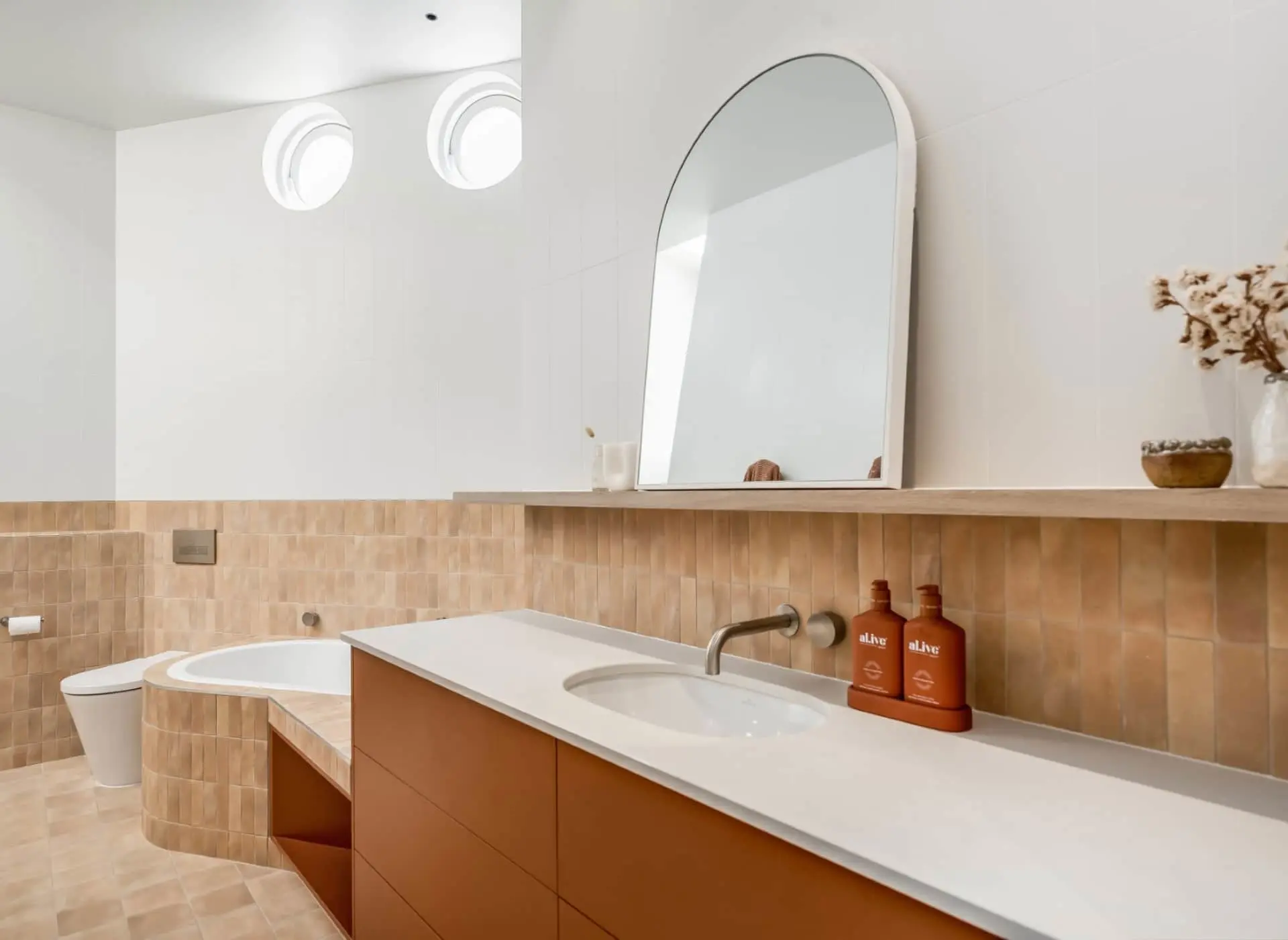 Modern bathroom with terracotta tiles, white countertop, arch mirror, rust vanity, and circular windows.