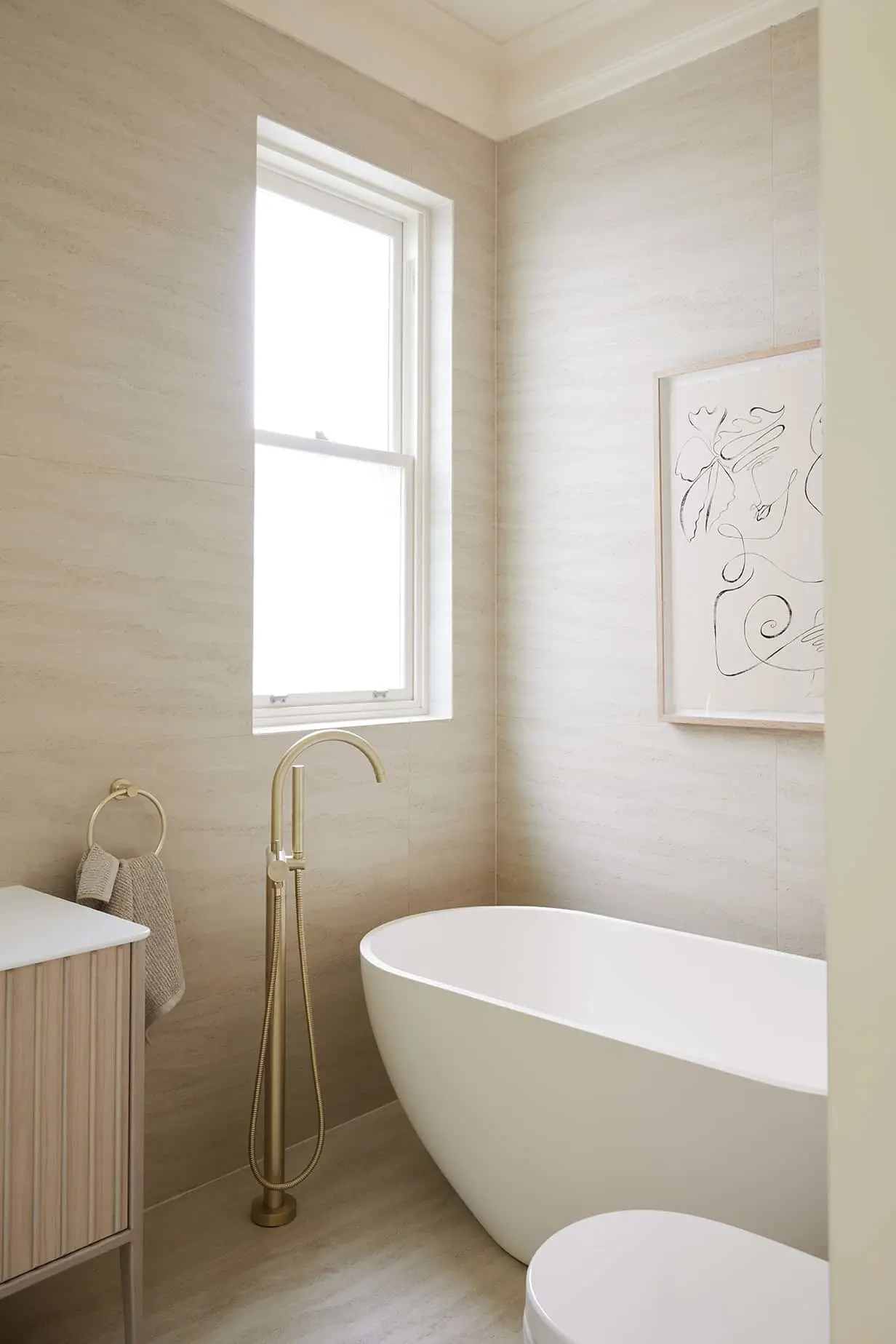 Modern bathroom with white freestanding tub, brass faucet, beige walls, window, and minimalist line art on wall.