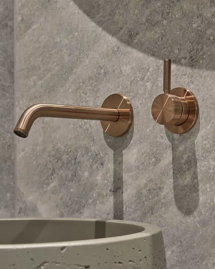 Bronze wall-mounted bathroom faucet with separate control knob installed on gray stone wall above concrete basin.