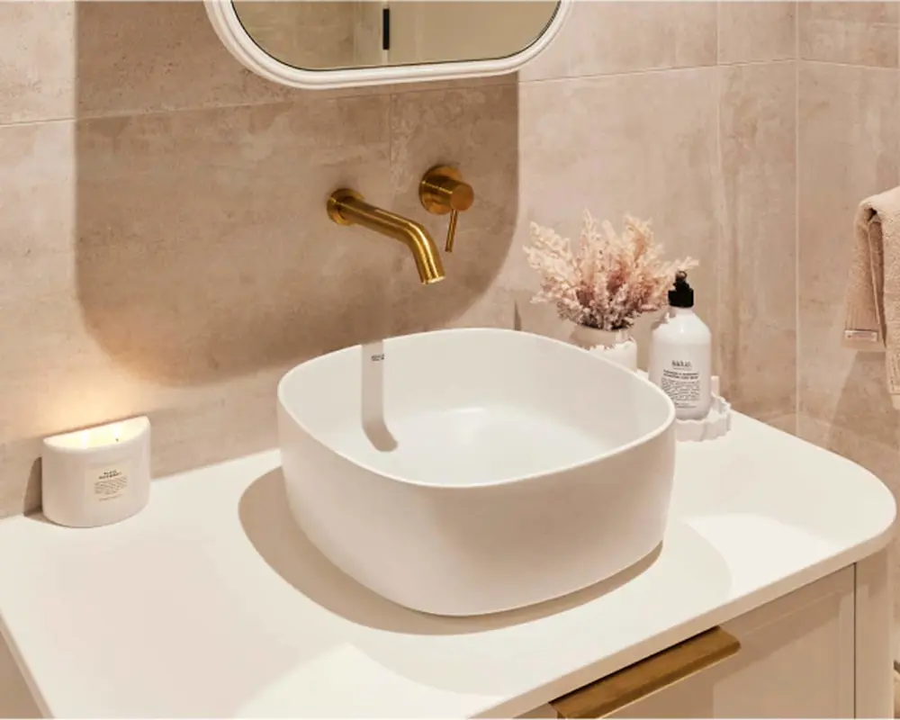 Modern bathroom with white vessel sink, brass wall-mounted faucet, beige tiled walls, and decorative dried flowers.