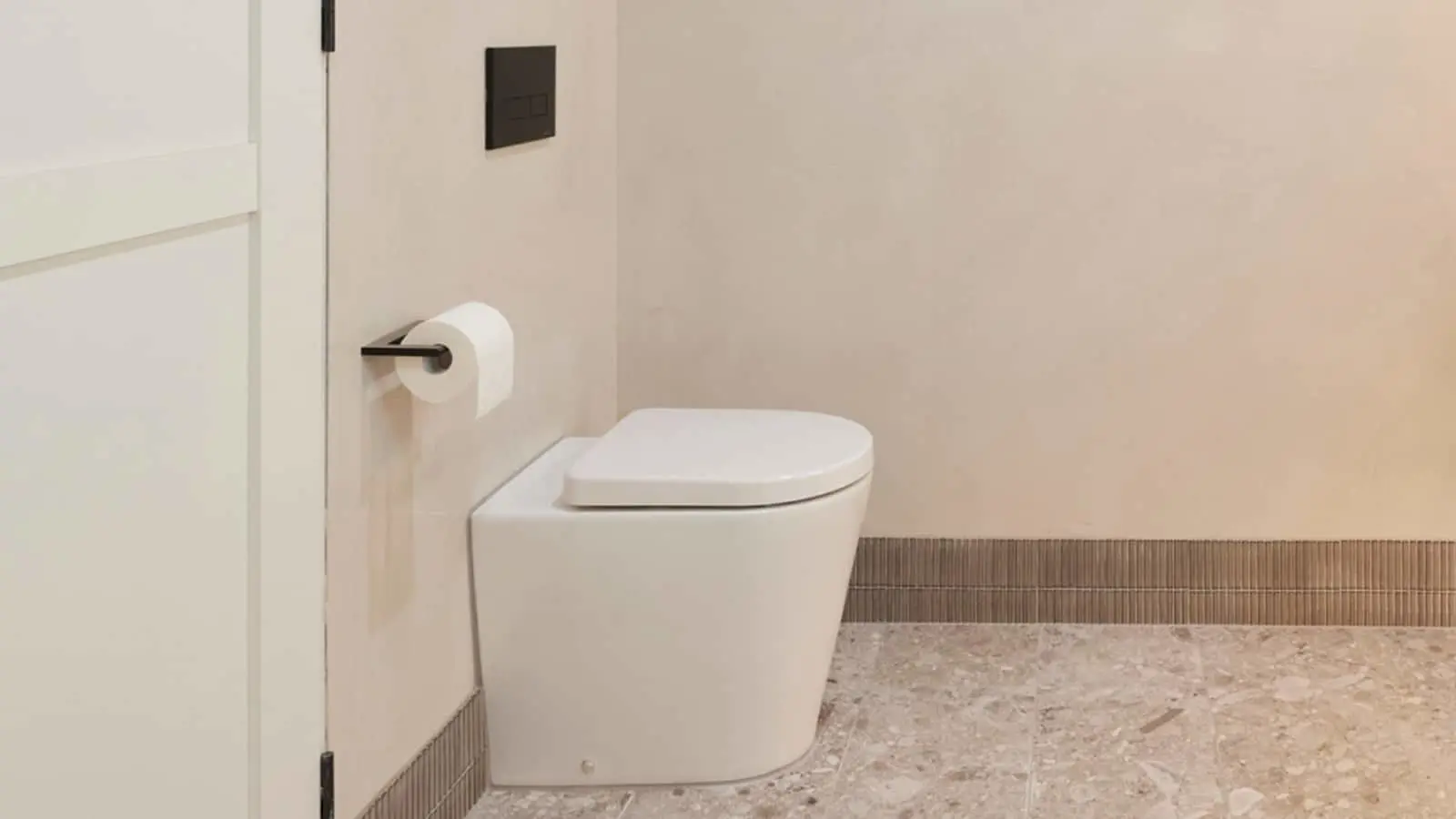 Modern white toilet with closed lid in minimalist bathroom with beige walls, stone flooring, and black flush panel.