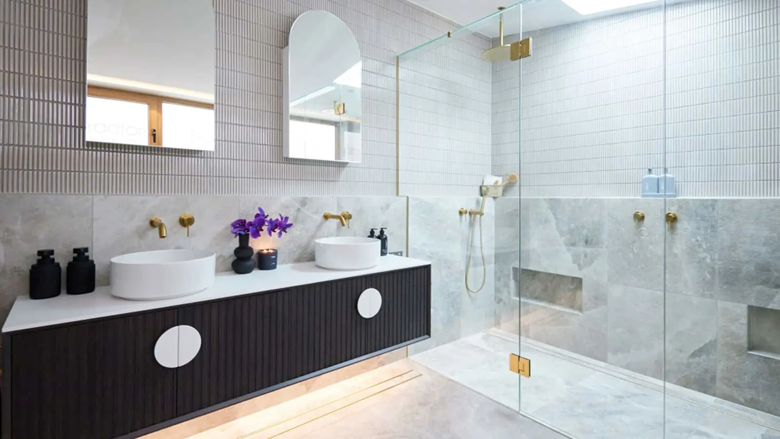 Modern bathroom with white vessel sinks, dark vanity, glass shower, marble walls, and purple flowers in a vase.