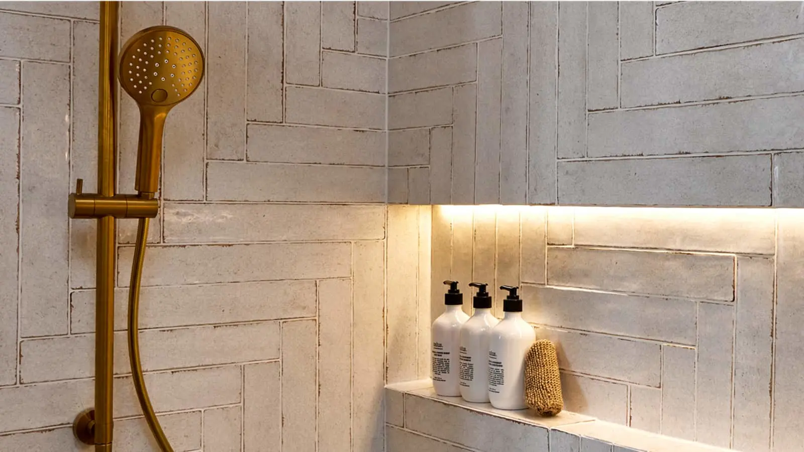 Modern bathroom with brass showerhead, white tiled walls, illuminated shelf holding three white pump bottles and loofah.