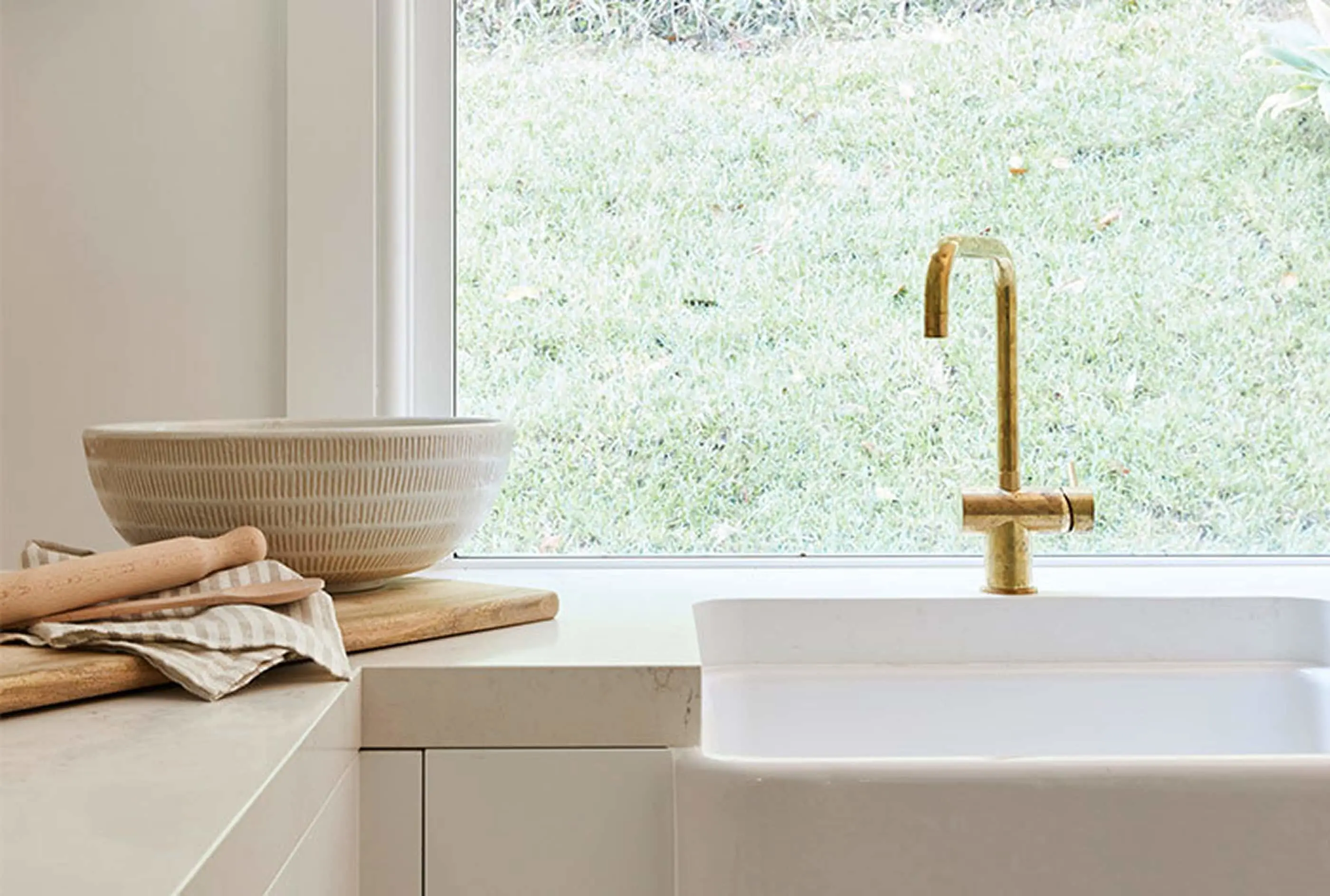 Modern kitchen with white sink, brass faucet, textured bowl, and wooden cutting boards beside a window overlooking grass.