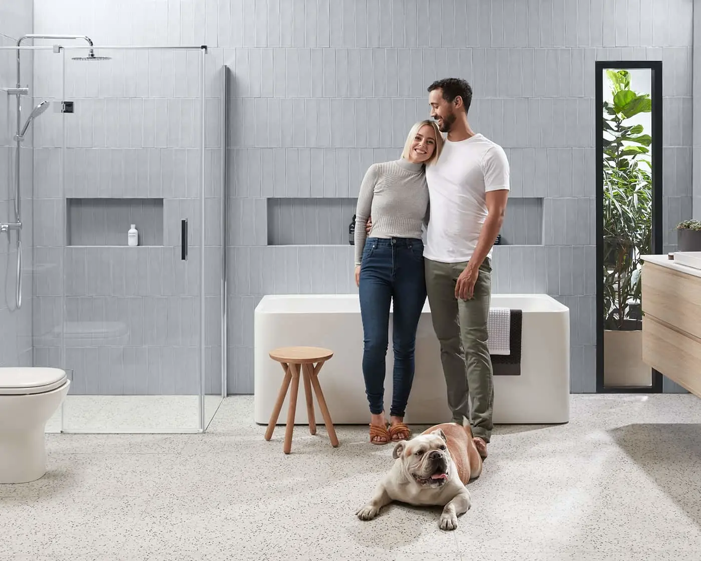 Couple standing in modern gray bathroom with white bathtub, glass shower, wooden stool, and bulldog lying on floor.