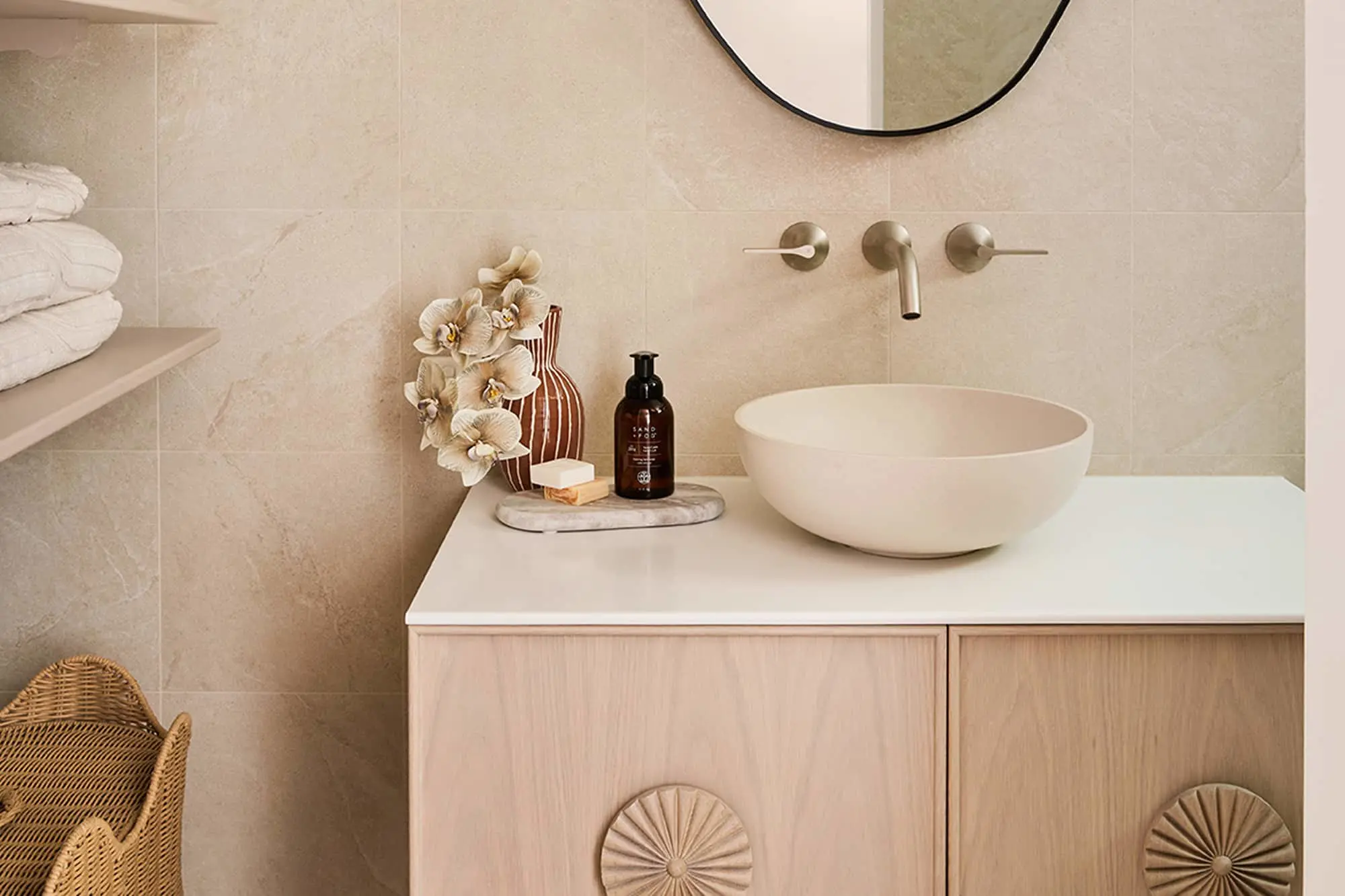 Neutral bathroom vanity with bowl sink, wall-mounted faucet, wood cabinet, round mirror, flowers, and soap bottle.