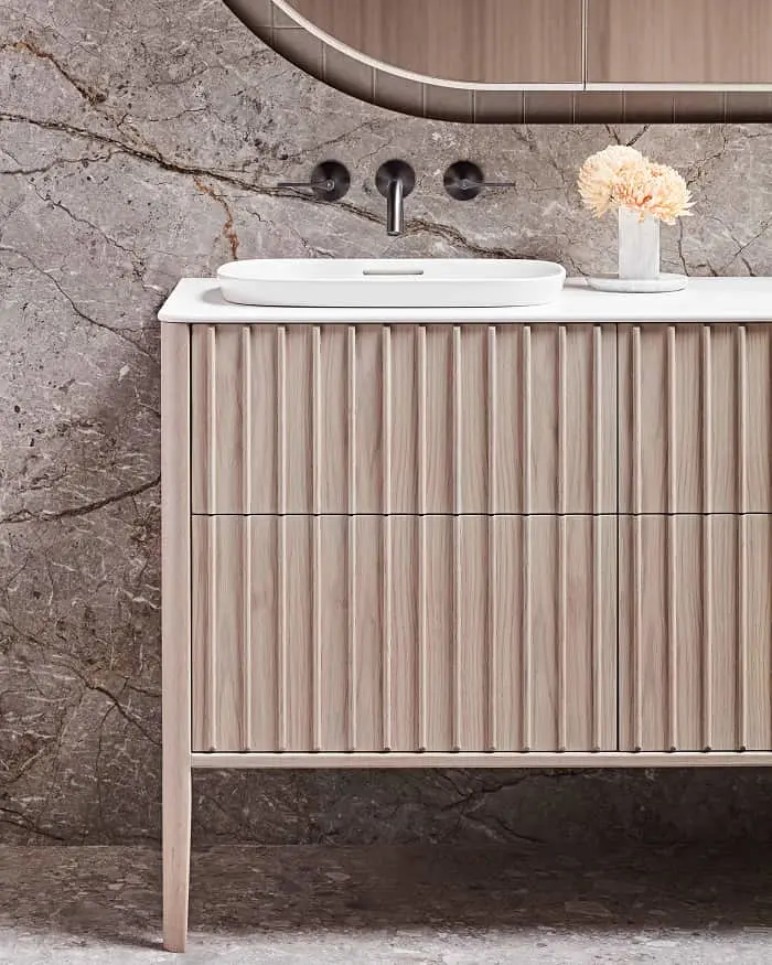 Modern bathroom vanity with light wood cabinetry, marble wall, white sink, wall-mounted faucet, and small flower vase.