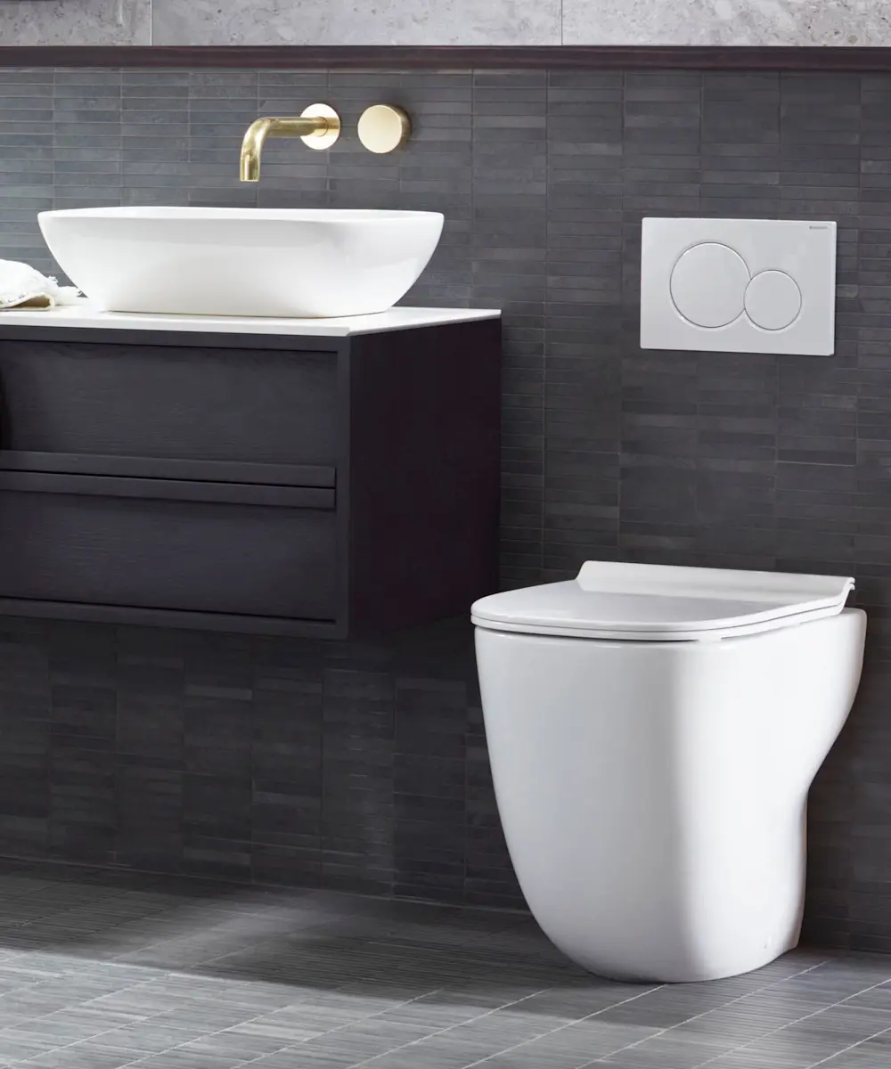 Modern bathroom with white vessel sink on dark vanity, wall-mounted toilet, brass faucet, and gray tile walls.